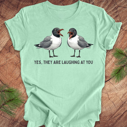 Mint green t-shirt with two laughing gulls and text, "yes, they are laughing at you" on a wood background.