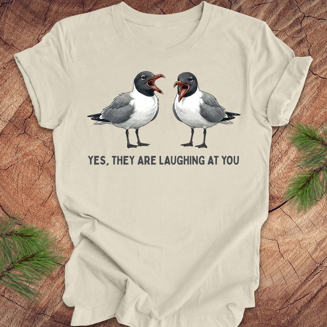 Beige t-shirt with two laughing gulls and text, "yes, they are laughing at you" on a wood background.