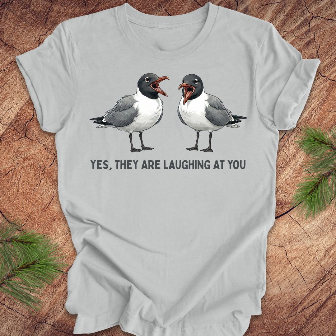 Gray t-shirt with two laughing gulls and text, "yes, they are laughing at you" on a wood background.