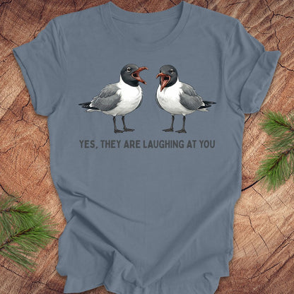 Blue t-shirt with two laughing gulls and text, "yes, they are laughing at you" on a wood background.