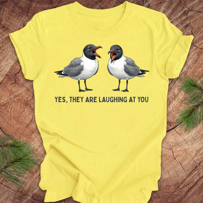 Yellow t-shirt with two laughing gulls and text, "yes, they are laughing at you" on a wood background.
