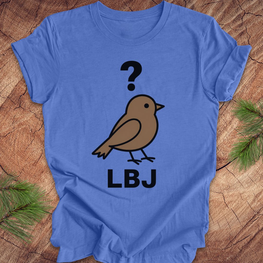 A blue short-sleeve t-shirt laid flat on a rustic wood surface with pine branches at the corners. The shirt features a graphic of a brown bird with a black question mark above it and the letters 'LBJ' printed below in black.
