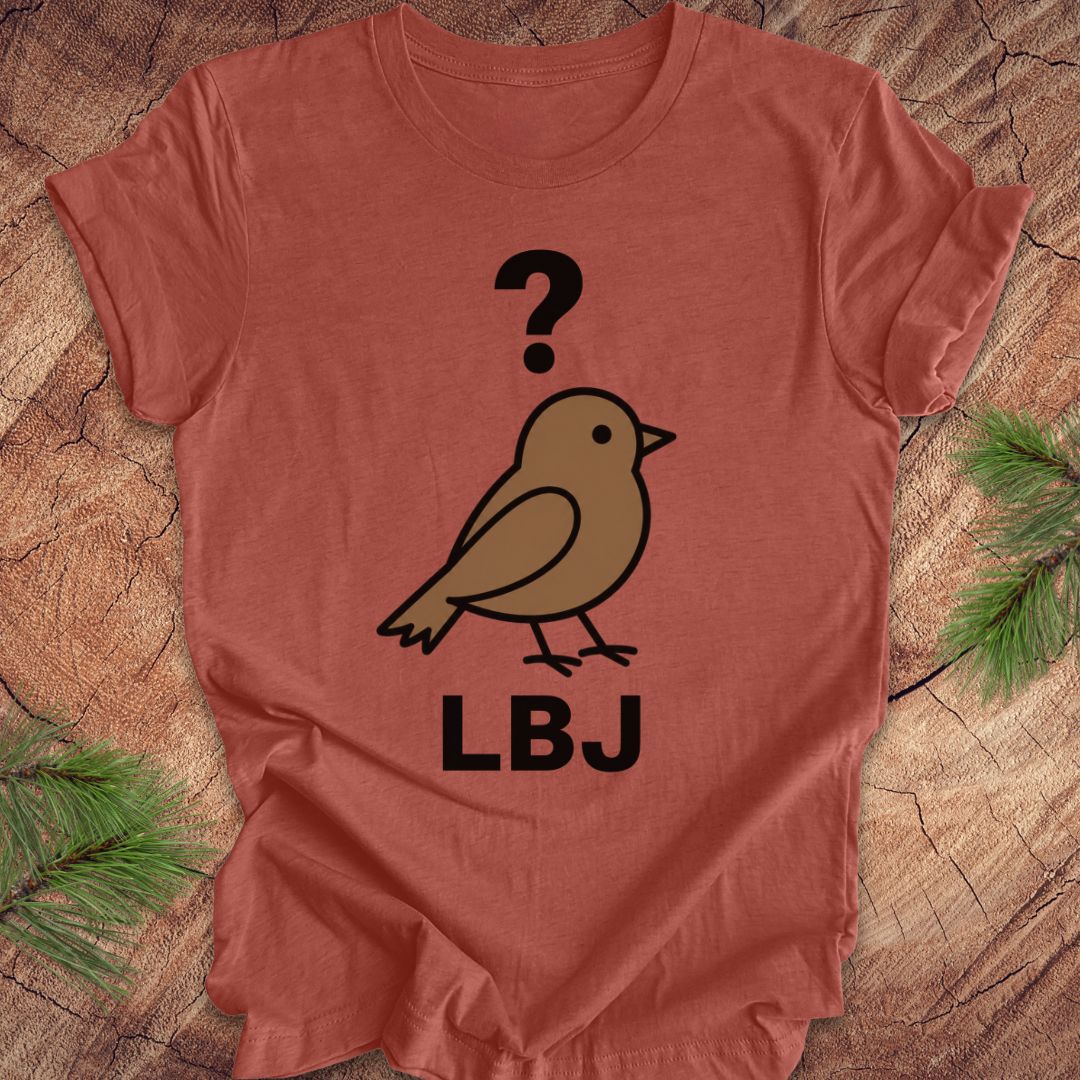 A terracotta-colored short-sleeve t-shirt laid flat on a rustic wood background with pine branches in the corners. The shirt features a graphic of a brown bird with a black question mark above it and the letters 'LBJ' printed below in black.