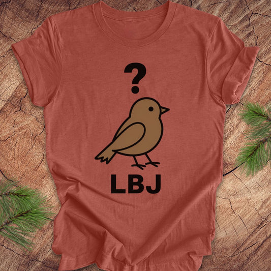 A terracotta-colored short-sleeve t-shirt laid flat on a rustic wood background with pine branches in the corners. The shirt features a graphic of a brown bird with a black question mark above it and the letters 'LBJ' printed below in black.
