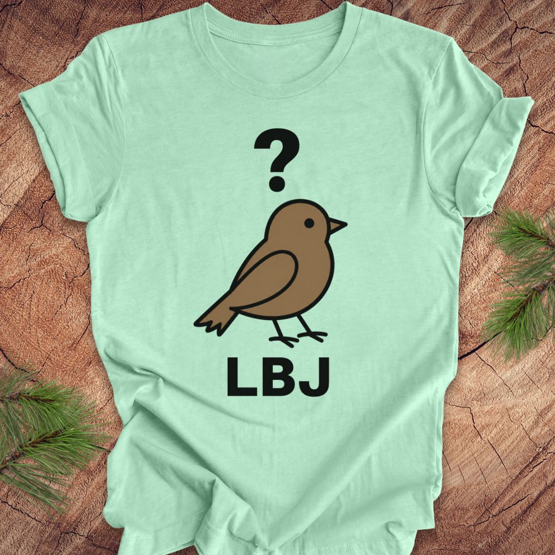 A mint green short-sleeve t-shirt laid flat on a rustic wood background with pine branches in the corners. The shirt features a graphic of a brown bird with a black question mark above it and the letters 'LBJ' printed below in black.