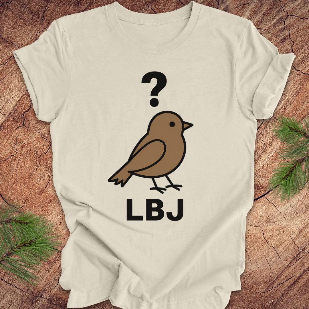 A beige short-sleeve t-shirt laid flat on a rustic wood background with pine branches in the corners. The shirt features a graphic of a brown bird with a black question mark above it and the letters 'LBJ' below in black.