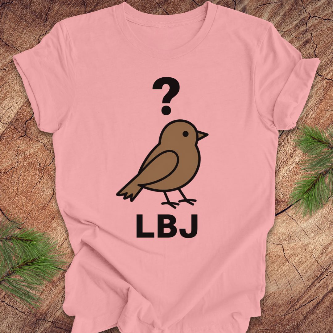 A light pink short-sleeve t-shirt laid flat on a rustic wood background with pine branches in the corners. The shirt features a graphic of a brown bird with a black question mark above it and the letters 'LBJ' printed below in black.