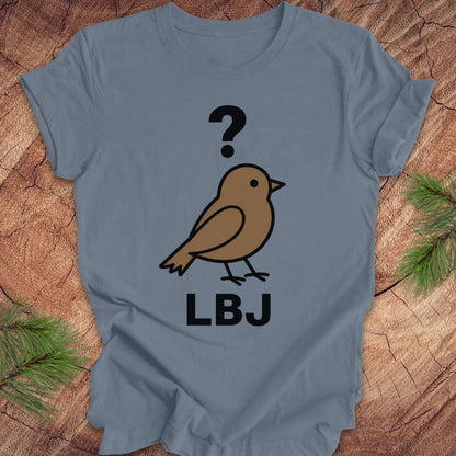 A slate blue short-sleeve t-shirt laid flat on a rustic wood background with pine branches at the corners. The shirt features a graphic of a brown bird with a black question mark above it and the letters 'LBJ' printed below in black.
