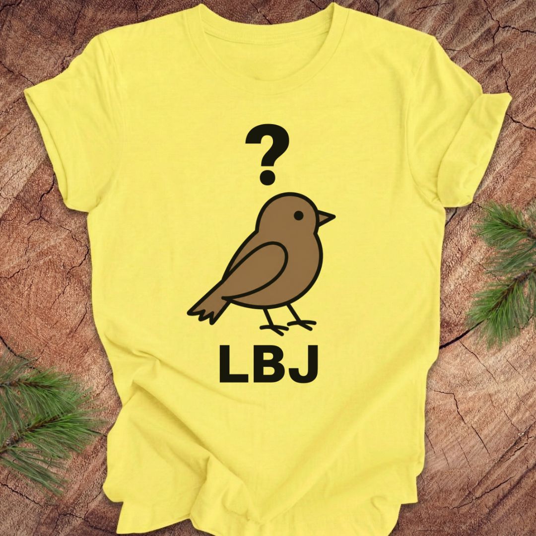A bright yellow short-sleeve T-shirt laid flat on a rustic wood surface with pine branches at the corners. The shirt has a printed graphic of a brown bird with a black question mark above it and the letters 'LBJ' below in black.