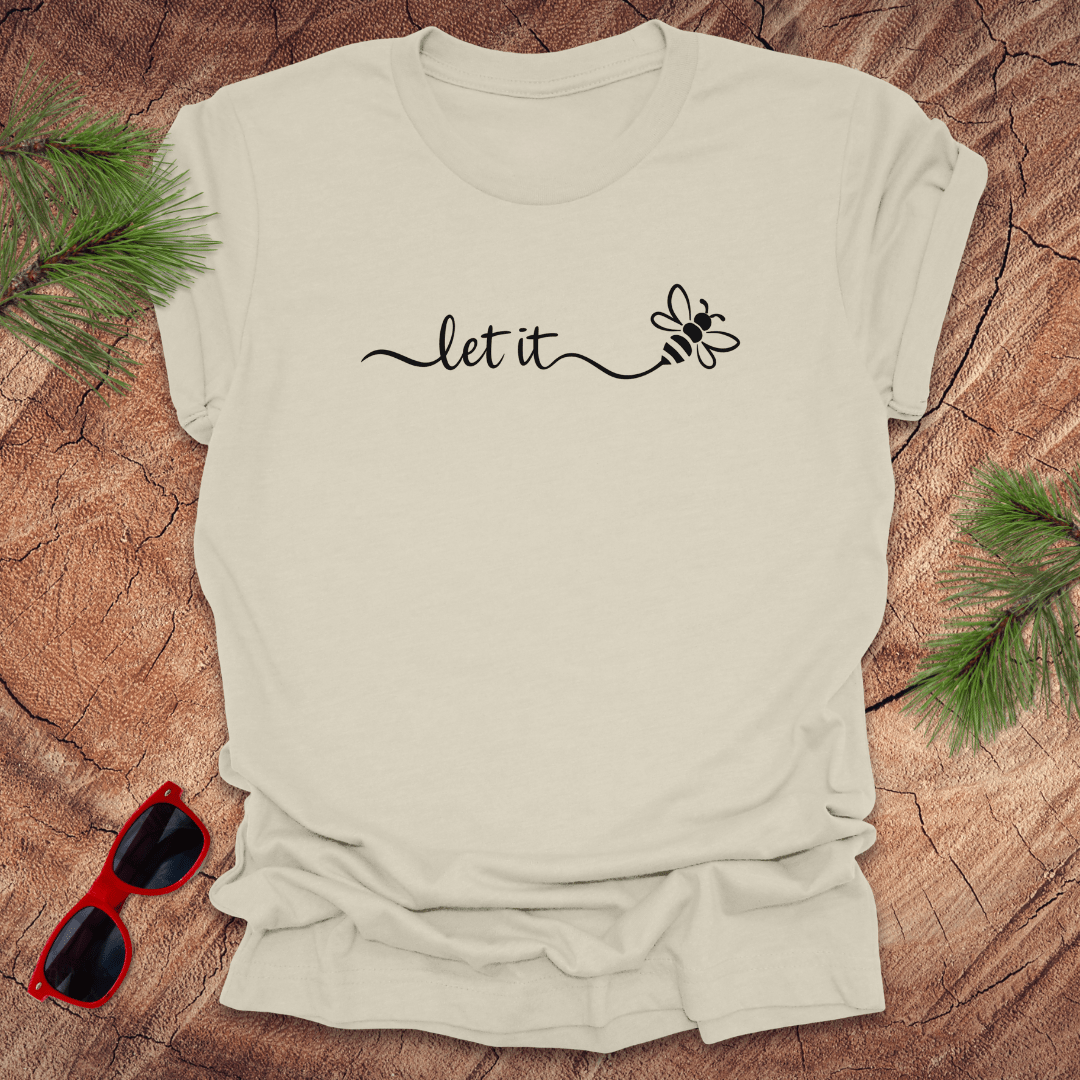Beige t-shirt with 'let it' and bee design on a wooden surface with sunglasses and greenery.