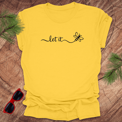 Yellow t-shirt with 'let it' and bee design on a wooden surface with sunglasses and greenery.