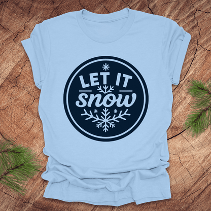 Light blue t-shirt with 'Let It Snow' design on a wooden surface with pine branches.