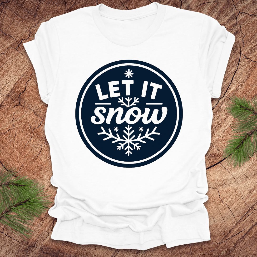 White t-shirt with 'Let It Snow' design on a wooden surface with pine branches.