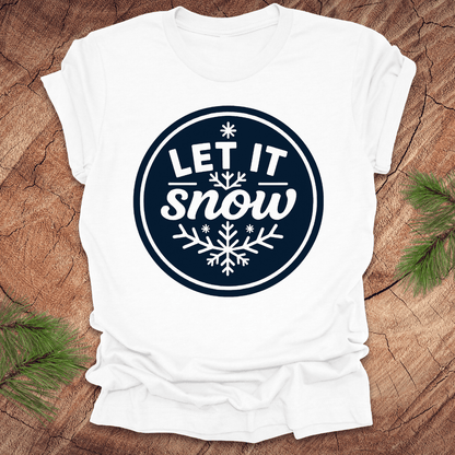 White t-shirt with 'Let It Snow' design on a wooden surface with pine branches.