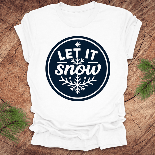 White t-shirt with 'Let It Snow' design on a wooden surface with pine branches.