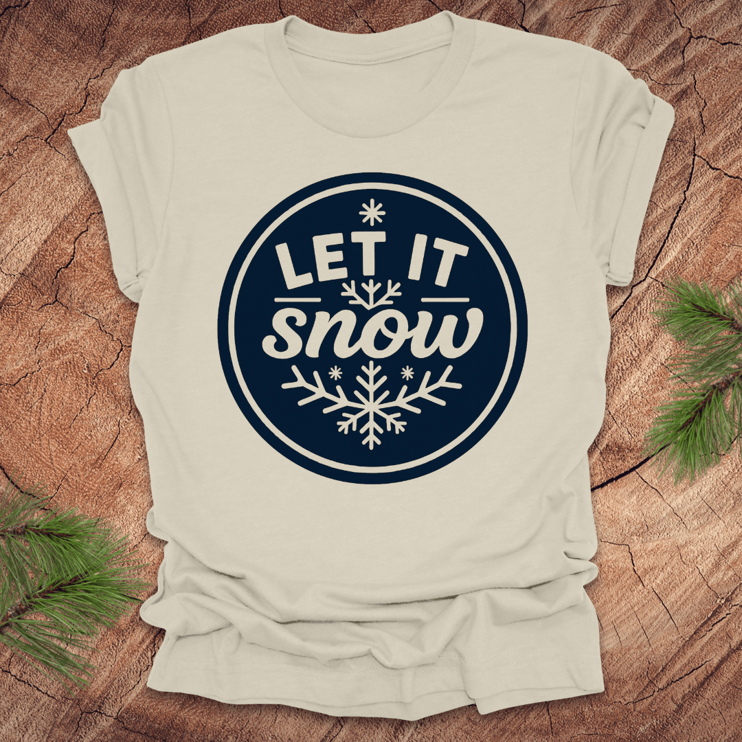Beige t-shirt with 'Let It Snow' design on a wooden surface with pine branches.
