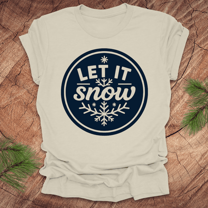 Beige t-shirt with 'Let It Snow' design on a wooden surface with pine branches.