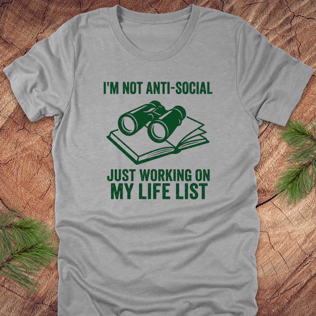 Grey t-shirt with green text 'i'm not anti-social just working on my life list' and binoculars graphic on a wooden surface