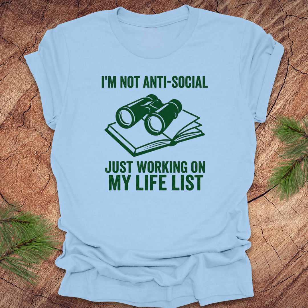 Baby blue t-shirt with green text 'i'm not anti-social just working on my life list' and binoculars graphic on a wooden surface