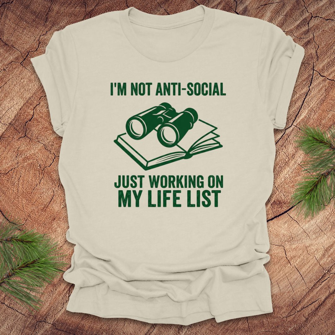 Beige t-shirt with green text 'i'm not anti-social just working on my life list' and binoculars graphic on a wooden surface