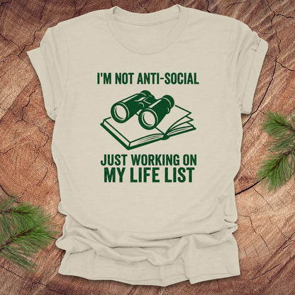 Beige t-shirt with green text 'i'm not anti-social just working on my life list' and binoculars graphic on a wooden surface