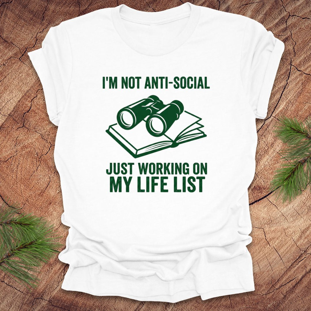 White t-shirt with green text 'i'm not anti-social just working on my life list' and binoculars graphic on a wooden surface
