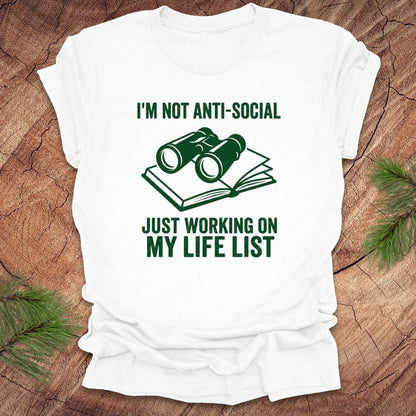 White t-shirt with green text 'i'm not anti-social just working on my life list' and binoculars graphic on a wooden surface