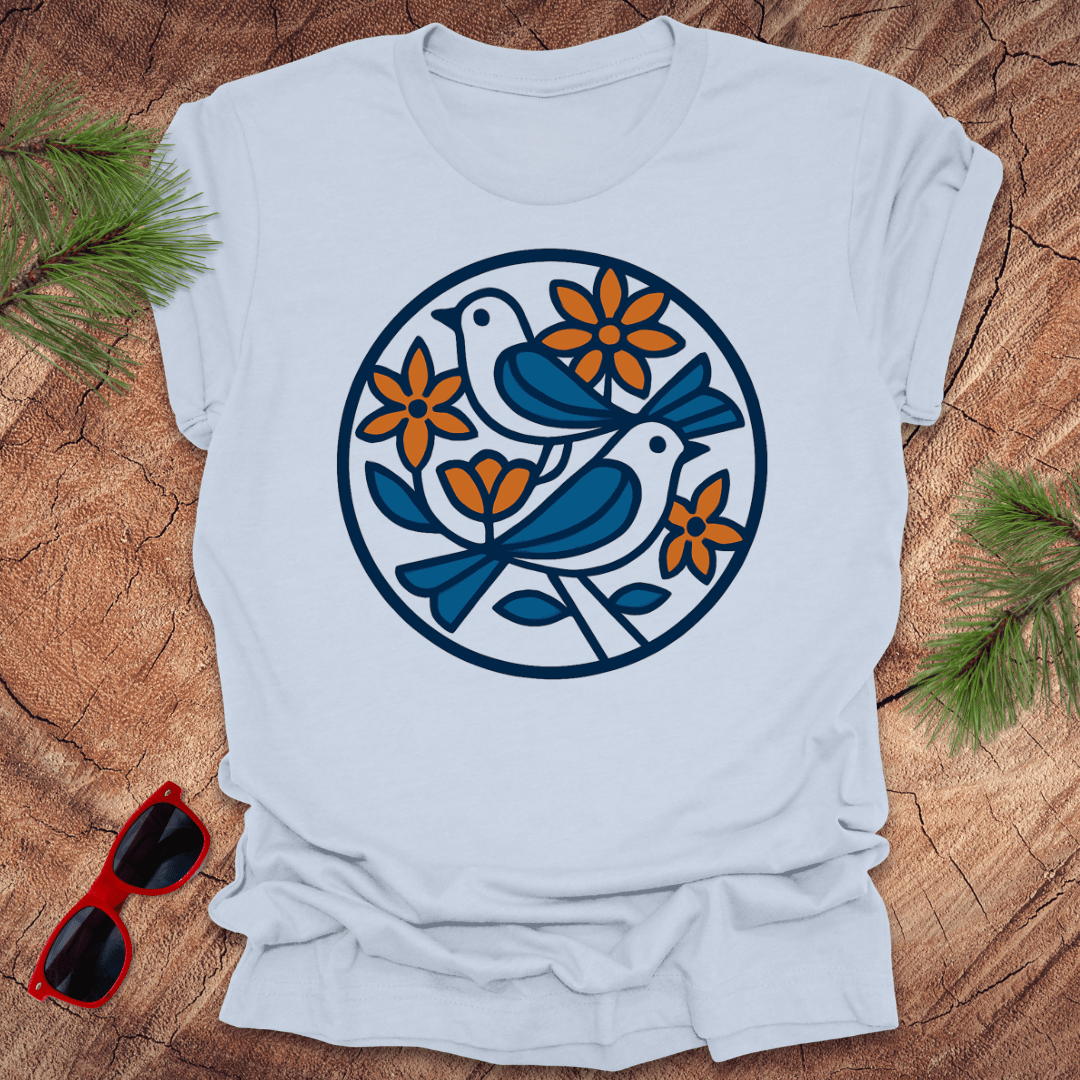 Light blue t-shirt with blue and orange bird and flower design on a wooden surface with sunglasses and greenery.