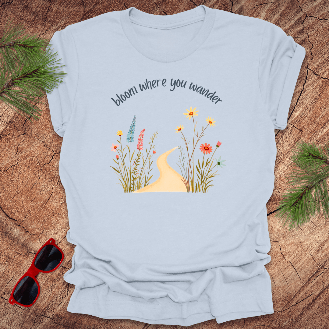blue t-shirt with a graphic that includes a floral design and the text 'bloom where you wander', accompanied by  flowers on a wooden background with pine needles and red sunglasses to the side.