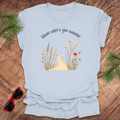 blue t-shirt with a graphic that includes a floral design and the text 'bloom where you wander', accompanied by  flowers on a wooden background with pine needles and red sunglasses to the side.