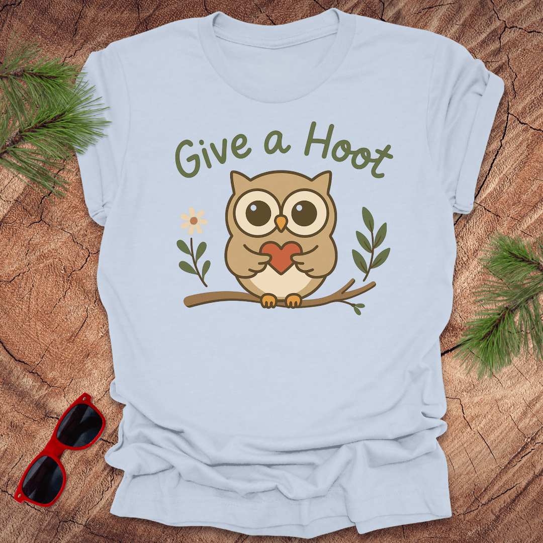 Light blue t-shirt with an owl graphic and 'Give a Hoot' text on a wooden surface with sunglasses and greenery.
