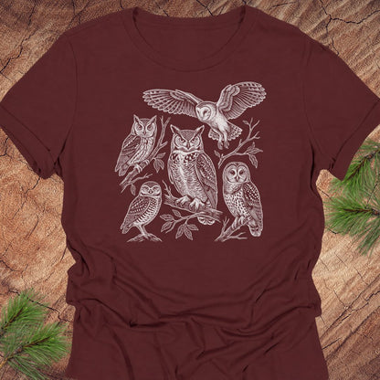 A dark brown short-sleeve t-shirt with a detailed white line art graphic of five owls perched on branches, laid flat on a textured wood background with pine needles in the corners.