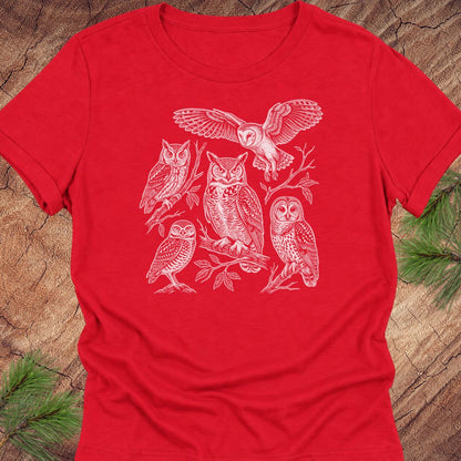 A red short-sleeve crewneck t-shirt featuring a white line art graphic of five different owl species, laid flat on a rustic wood background with pine needles visible in the corners.