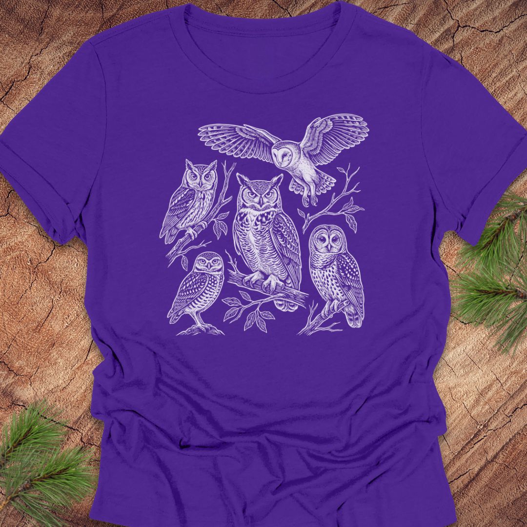A purple short-sleeve crewneck t-shirt featuring a white line art graphic of five different owl species, laid flat on a rustic wood background with pine needles in the corners.