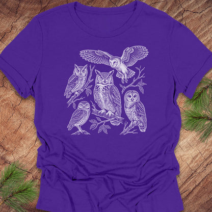 A purple short-sleeve crewneck t-shirt featuring a white line art graphic of five different owl species, laid flat on a rustic wood background with pine needles in the corners.