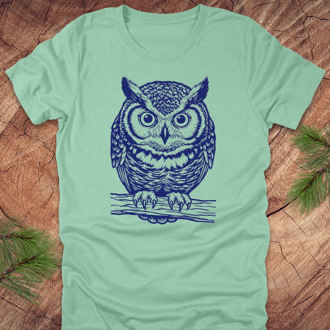 Mint green t-shirt with a linocut style owl, in a blue ink, sitting on a branch, on a wood background.