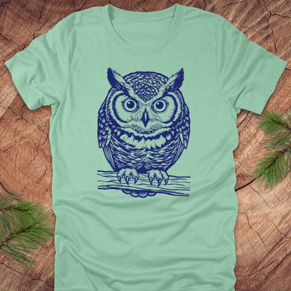 Mint green t-shirt with a linocut style owl, in a blue ink, sitting on a branch, on a wood background.