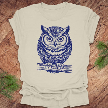 Beige t-shirt with a linocut style owl, in a blue ink, sitting on a branch, on a wood background.