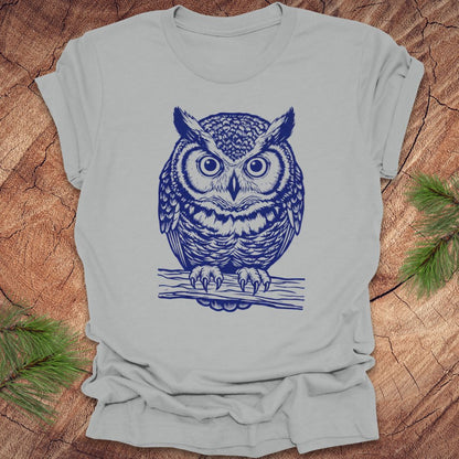 Silver t-shirt with a linocut style owl, in a blue ink, sitting on a branch, on a wood background.