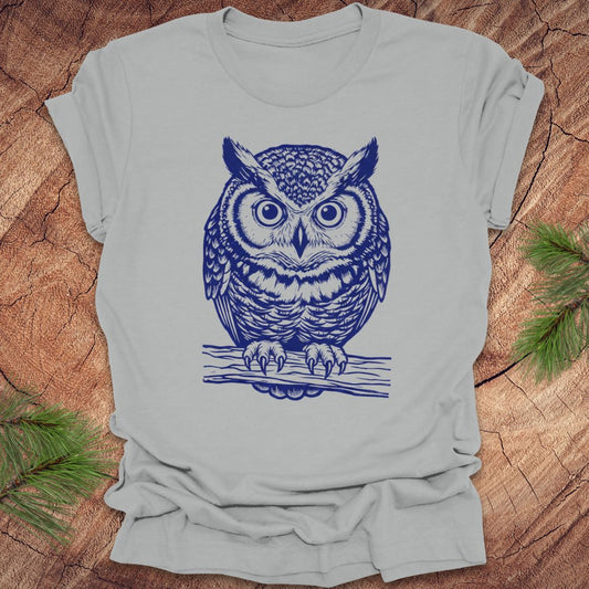 Silver t-shirt with a linocut style owl, in a blue ink, sitting on a branch, on a wood background.