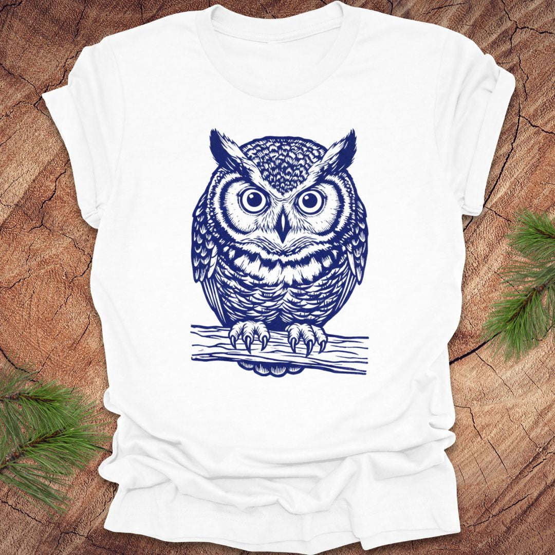 White t-shirt with a linocut style owl, in a blue ink, sitting on a branch, on a wood background.