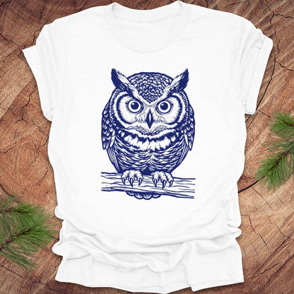 White t-shirt with a linocut style owl, in a blue ink, sitting on a branch, on a wood background.