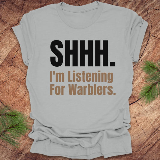 Gray t-shirt with 'SHHH. I'm Listening For Warblers.' text on a wooden surface with greenery.