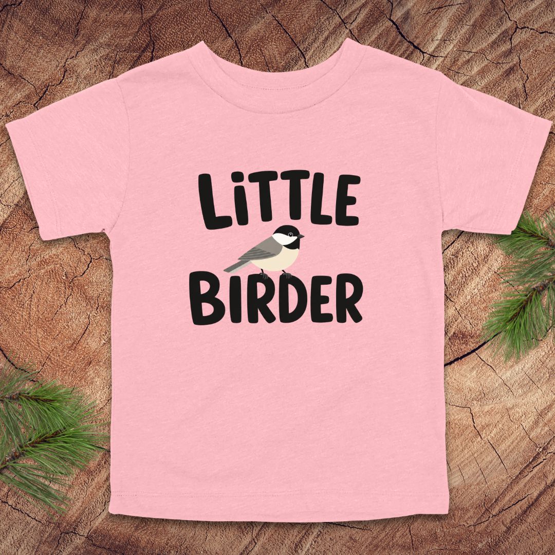 Pink t-shirt with 'Little Birder' text and a bird illustration on a wooden surface.
