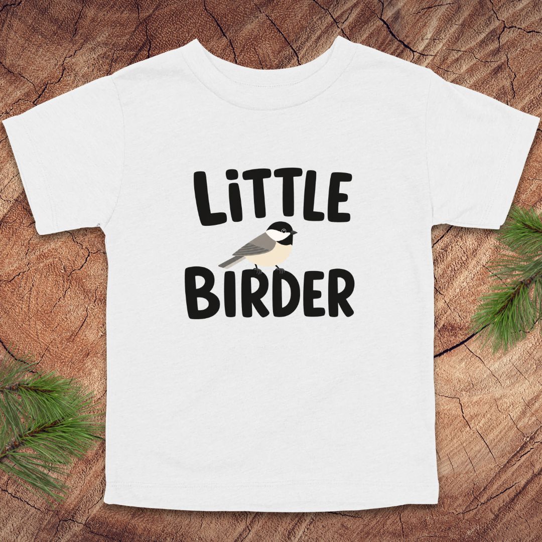 White t-shirt with 'Little Birder' text and a bird illustration on a wooden surface.