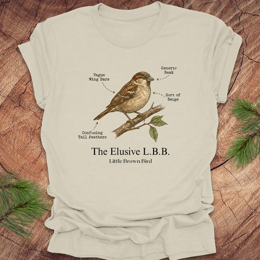Beige t-shirt with a bird illustration and text 'the elusive L.B.B" on a wooden surface