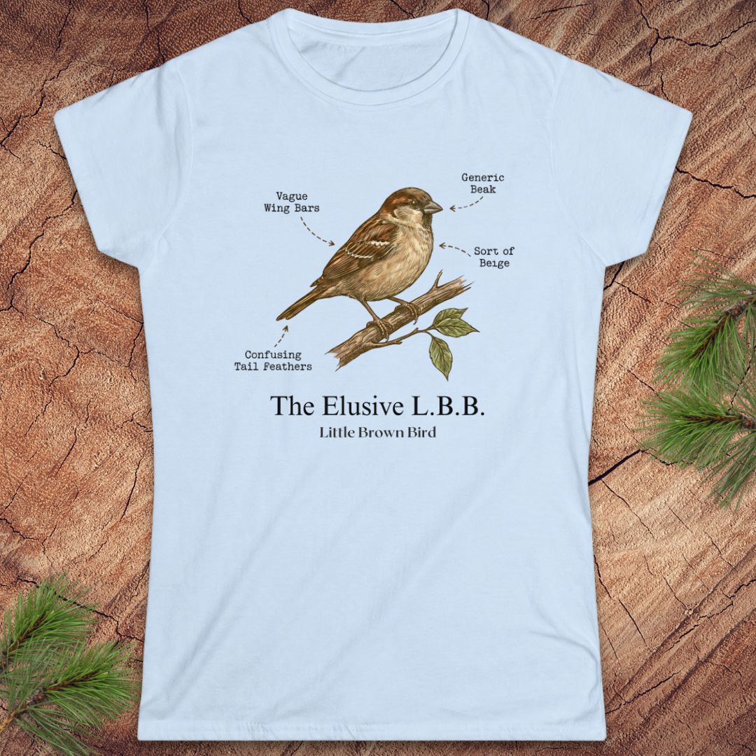 A light blue women's short-sleeve t-shirt with a vintage-style illustration of a little brown bird on a branch, labeled with humorous text, displayed flat on a rustic wood background with pine needles.