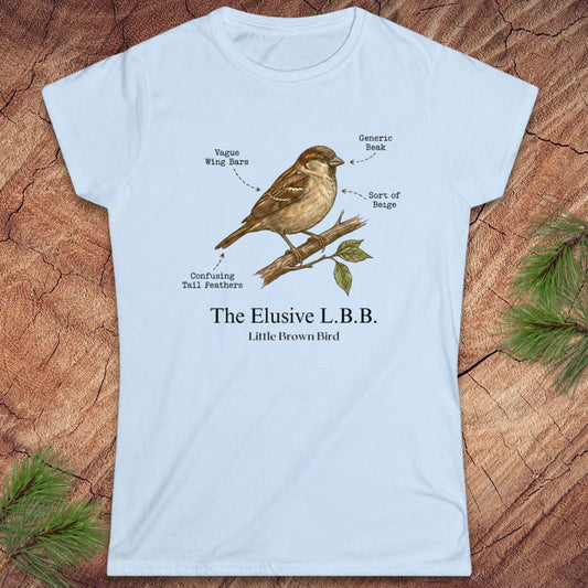 A light blue women's short-sleeve t-shirt with a vintage-style illustration of a little brown bird on a branch, labeled with humorous text, displayed flat on a rustic wood background with pine needles.