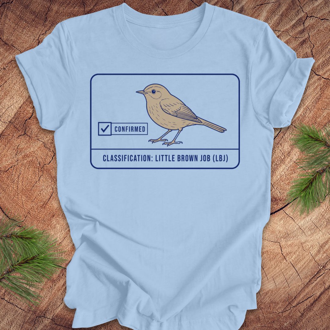 A light blue short-sleeve t-shirt laid flat on a rustic wood surface with pine branches in the corners. The shirt features a graphic of a little brown bird with a 'Confirmed' badge and the text 'CLASSIFICATION: LITTLE BROWN JOB (LBJ)'.