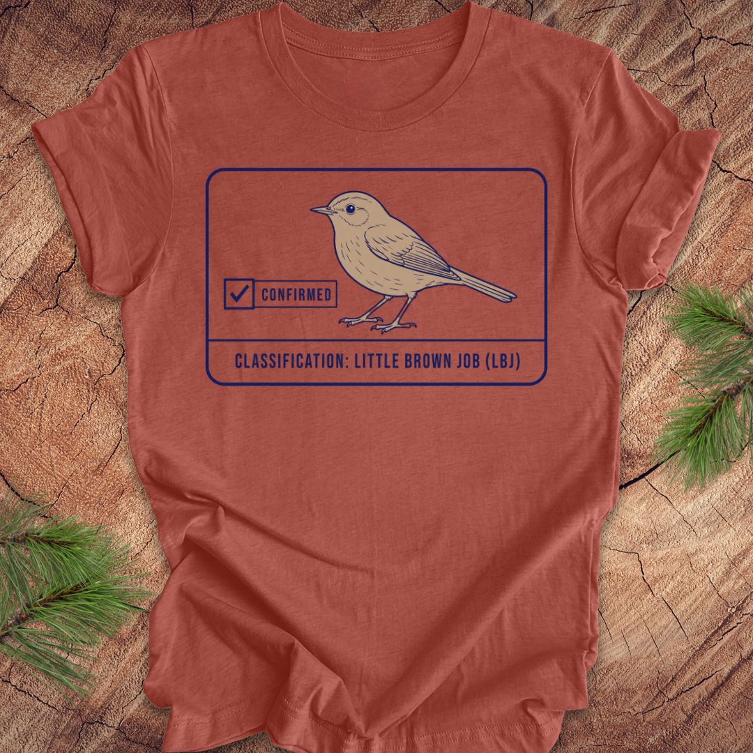 A rust-orange crewneck t-shirt featuring a graphic of a little brown bird with a 'Confirmed' stamp and the text 'CLASSIFICATION: LITTLE BROWN JOB (LB)'. The shirt is laid flat on a rustic wood background with pine needles in the corners.